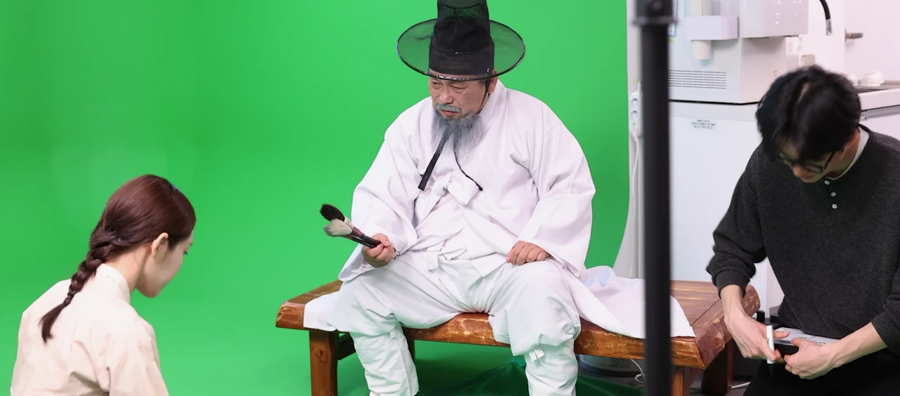 An actor in Joseon-era costume holding a real brush on a greenscreen set